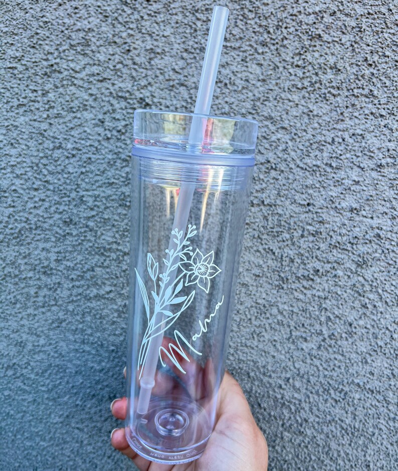 May include: Clear plastic tumbler with a white floral design and the word "Momma" written in cursive. The tumbler has a straw.