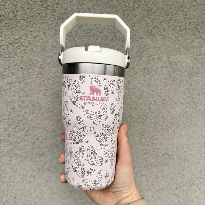 May include: A pink stainless steel travel mug with a white lid and handle. The mug has a black and white butterfly pattern and the word "STANLEY" printed on the side.