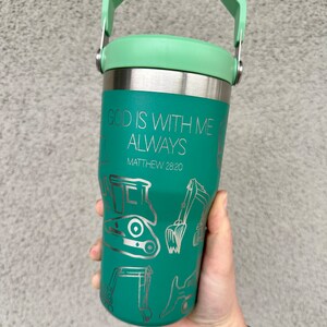 May include: Teal stainless steel travel mug with a green lid and a carrying handle. The mug has a silver inscription that reads "God is with me always Matthew 28:20" and a design of construction equipment.