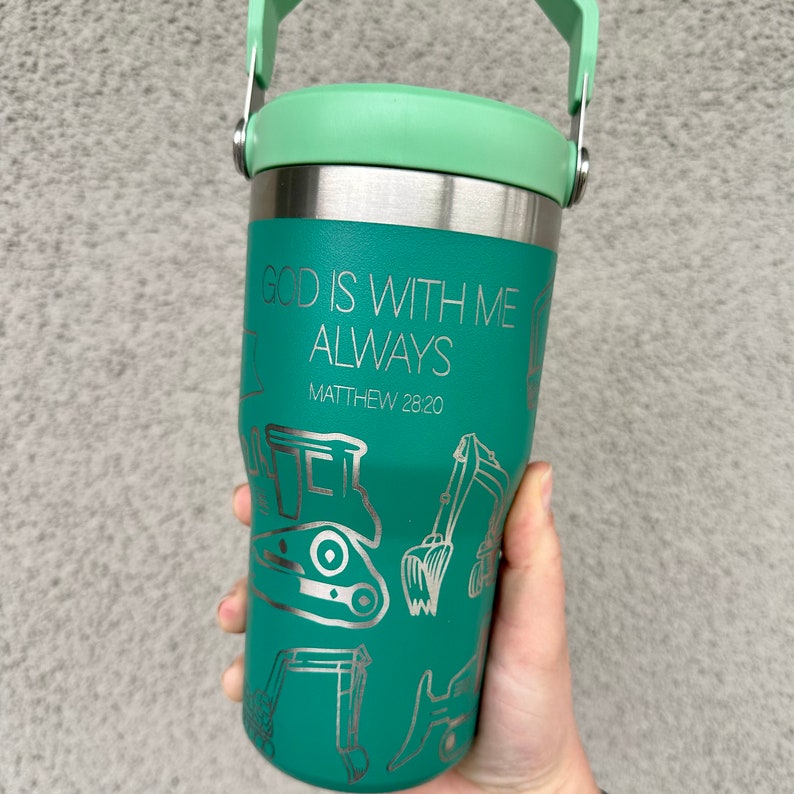 May include: Teal stainless steel travel mug with a green lid and a carrying handle. The mug has a silver inscription that reads "God is with me always Matthew 28:20" and a design of construction equipment.