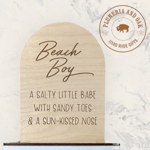 May include: A wooden sign with a rounded top, featuring the text "Beach Boy" in a cursive font. The sign also includes the text "A salty little babe with sandy toes & a sun-kissed nose". The sign is made by Plumeria and Oak, a company that makes handmade gifts.