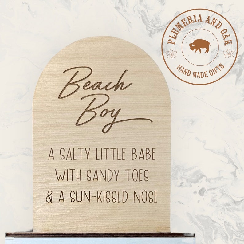 May include: A wooden sign with a rounded top, featuring the text "Beach Boy" in a cursive font. The sign also includes the text "A salty little babe with sandy toes & a sun-kissed nose". The sign is made by Plumeria and Oak, a company that makes handmade gifts.