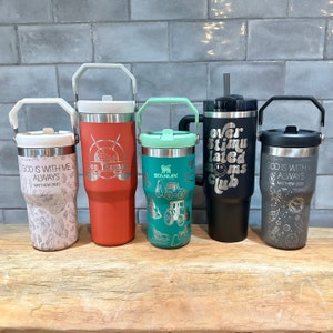 May include: Five stainless steel tumblers with various designs and colors. The tumblers feature different engraved sayings, including "God is with me always", "On Thomas", "Over Stimulated Moms Club", and "Stanley".