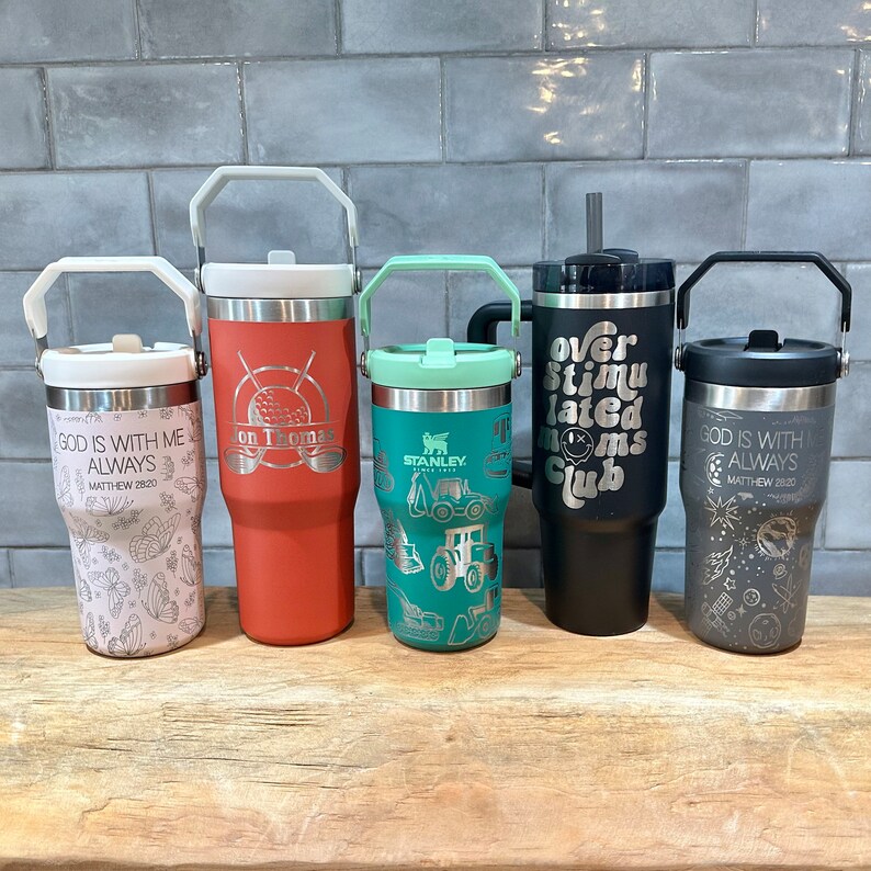May include: Five stainless steel tumblers with various designs and colors. The tumblers feature different engraved sayings, including "God is with me always", "On Thomas", "Over Stimulated Moms Club", and "Stanley".