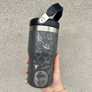 May include: A gray stainless steel travel mug with a black lid and handle. The mug has a space-themed design with stars, planets, and a rocket ship.
