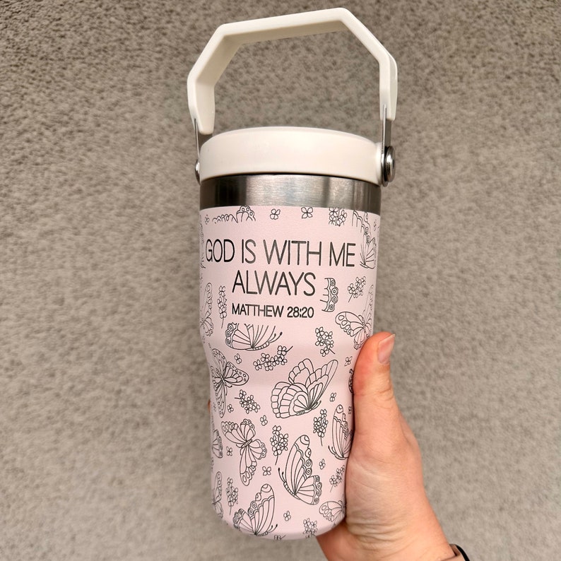 May include: A pink stainless steel travel mug with a white lid and a black handle. The mug has a black and white design of butterflies and flowers. The text "GOD IS WITH ME ALWAYS" and "MATTHEW 28:20" is printed on the mug.