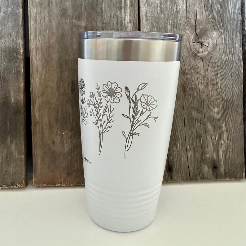 May include: White stainless steel tumbler with a floral design etched on the side. The design features a bouquet of flowers with stems and leaves.