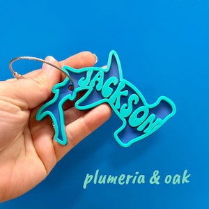 May include: A blue and teal luggage tag in the shape of a hammerhead shark, with the name "Jackson" in teal lettering. The tag has a silver metal loop for attachment and the text "plumeria & oak" in teal.