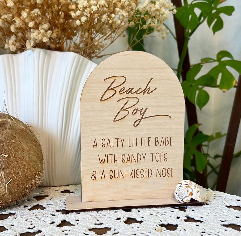 May include: Wooden sign with a rounded top, featuring the text "Beach Boy" in cursive script. Below the text, it reads "A salty little babe with sandy toes & a sun-kissed nose".
