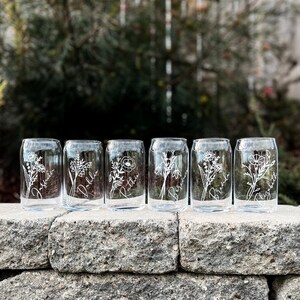 May include: Six clear glass tumblers with etched floral designs and names, including "Josephine", "Dahlia", "Ruthie", "Bertha", "Brenda", and "Charlotte", arranged on a stone wall.