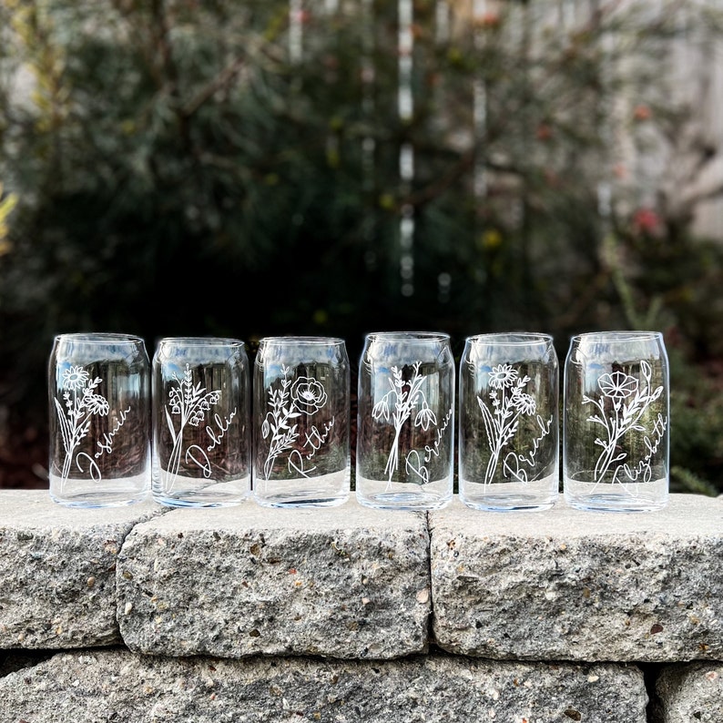 May include: Six clear glass tumblers with etched floral designs and names, including "Josephine", "Dahlia", "Ruthie", "Bertha", "Brenda", and "Charlotte", arranged on a stone wall.