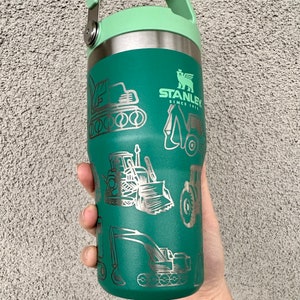 May include: Green stainless steel water bottle with a green lid and a handle. The bottle has a silver design of construction equipment and the text "STANLEY SINCE 1913".