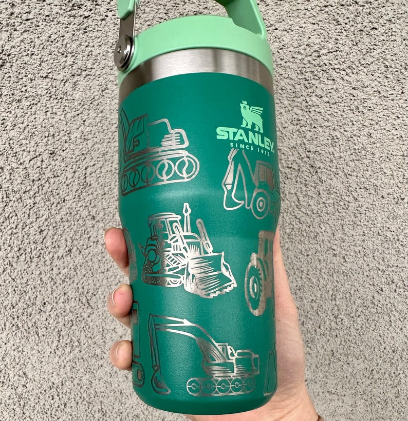 May include: Green stainless steel water bottle with a green lid and a handle. The bottle has a silver design of construction equipment and the text "STANLEY SINCE 1913".