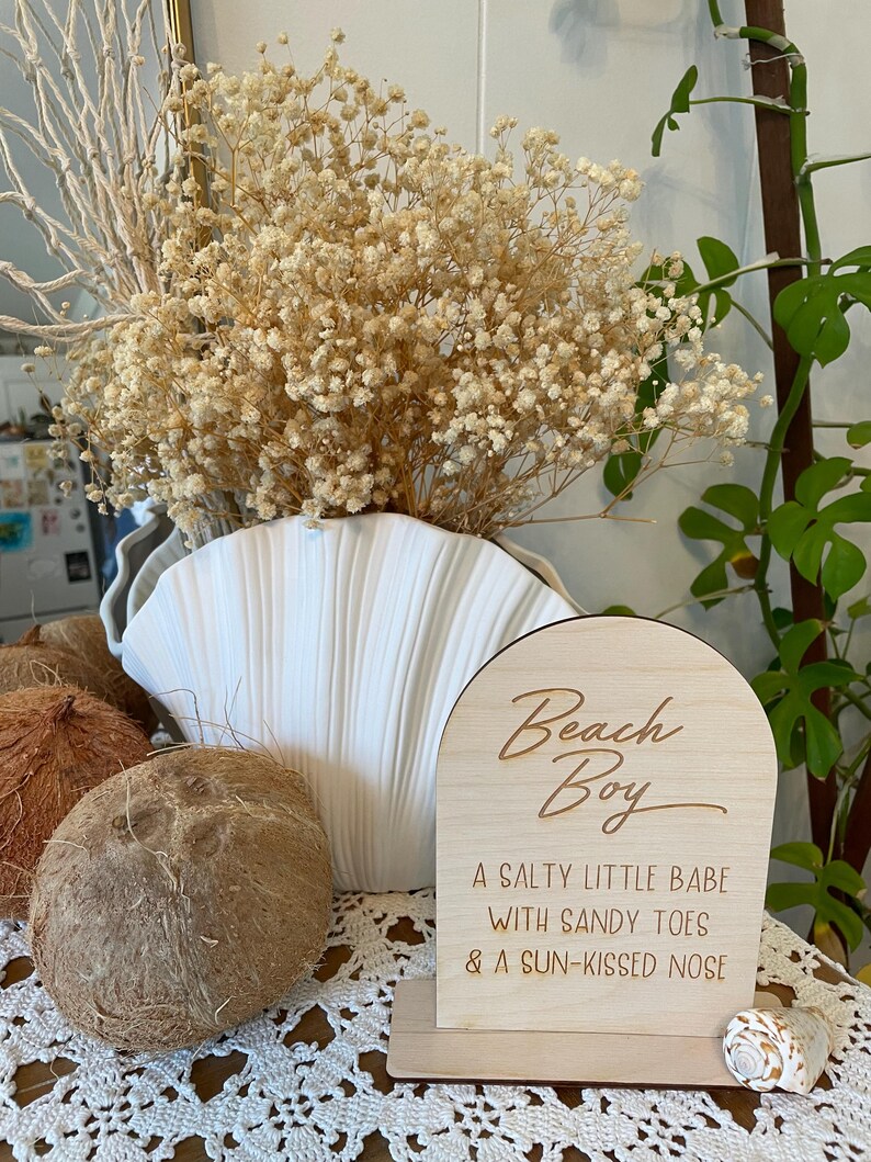 May include: Wooden sign with a rounded arch top, reading "Beach Boy" in cursive script. The sign has a light wood finish and the text is in a darker brown color. Below the text, it reads "A salty little babe with sandy toes & a sun-kissed nose".