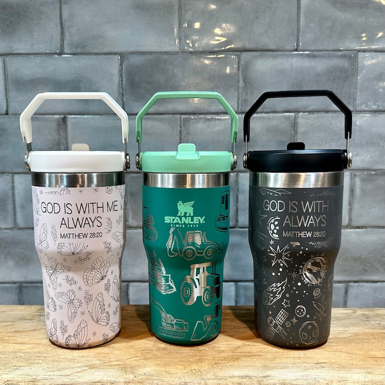 May include: Three stainless steel travel mugs with different designs. The first mug is white with a pink butterfly design and the text "God is with me always Matthew 28:20". The second mug is green with a construction equipment design and the Stanley logo. The third mug is gray with a space-themed design and the text "God is with me always Matthew 28:20".