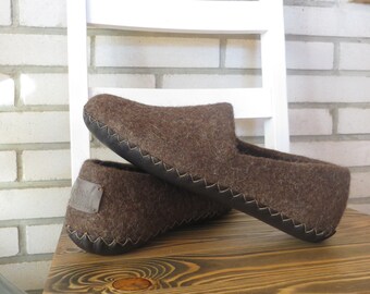Può includere: Un paio di pantofole in lana marrone con bordi e cuciture in pelle nera. Le pantofole sono su una superficie in legno.