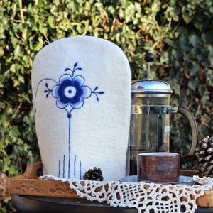Puede incluir: Un calienta-té de fieltro blanco con un diseño floral azul y blanco. El calienta-té está sobre una bandeja de madera con una cafetera de émbolo y una taza.