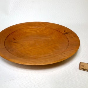 May include: A round wooden bowl with a natural wood finish. The bowl has a raised rim and is made of cherry wood.
