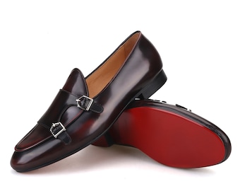 May include: A pair of burgundy leather double monk strap shoes with silver buckles. The shoes have a sleek design with a pointed toe and a red sole. The shoes are displayed on a white background.