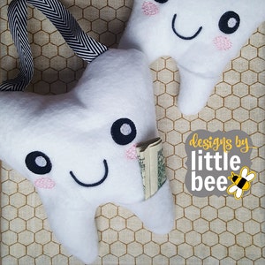 May include: Two white plush tooth pillows with embroidered smiling faces. One tooth pillow has a pocket with a rolled up dollar bill inside. The pillows are on a brown and white patterned background. The text "designs by little bee" is in a gray circle with a yellow bee.