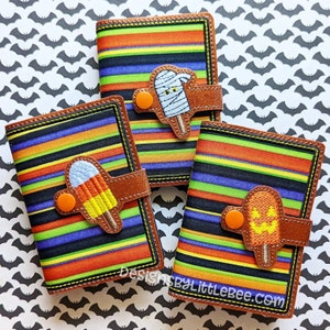 Puede incluir: Tres cuadernos con temática de Halloween con cubiertas a rayas coloridas y cierres de cuero marrón. Cada cuaderno presenta un diseño de paleta diferente: una momia, un caramelo de maíz y una calabaza. Fondo con siluetas de murciélagos.