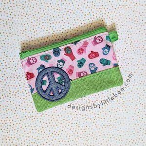May include: A rectangular zippered pouch featuring a light pink fabric with a repeating pattern of colorful mittens and paw prints. The bottom section is a textured green fabric, and a dark blue circular peace sign patch is stitched on the front.
