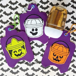 Puede incluir: Llaveros morados con temática de Halloween con diseños de cubos bordados. Un llavero presenta un cubo fantasma, otro un cubo de bruja y el tercero un cubo de calabaza. También hay un frasco transparente de desinfectante de manos con base blanca.