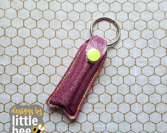 Puede incluir: Un porta bálsamo labial de purpurina rosa con un botón amarillo y un llavero plateado. El porta bálsamo está sobre un fondo blanco y dorado con patrón de panal.