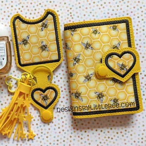May include: A yellow and black bee-themed key chain with a heart-shaped charm and a tassel. A matching yellow and black bee-themed wallet with a heart-shaped flap. The wallet has the text "designsbylittlebee.com" on the front.