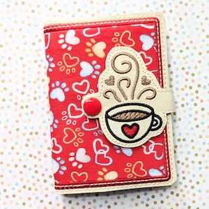 May include: A red fabric wallet with a white and red heart pattern. The wallet has a flap with a white button and a brown embroidered coffee cup with steam.