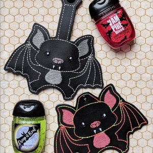 May include: Two black bat-shaped hand sanitizer holders with embroidered details. One holder has a black bat with white stitching and the other has a black bat with pink stitching and neon green accents. The holders are attached to small bottles of hand sanitizer. One bottle is green and says "Creep it Real" and the other is red and says "Jam Pire Blood".