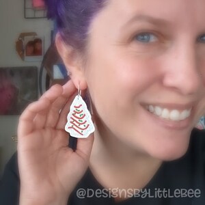 May include: White, tree-shaped earrings with red and green accents. The earrings are a festive design, perfect for the holiday season. The earrings are hanging from silver hooks.