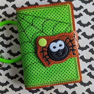 Puede incluir: Una cartera verde y negra con temática de Halloween con un diseño de telaraña. La cartera presenta una aplicación de araña marrón con una cara sonriente y un cierre de botón verde. Una pulsera verde está unida a la cartera.
