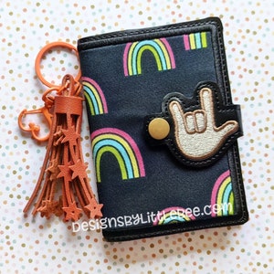 Op de afbeelding: Een zwarte stoffen portemonnee met een regenboogpatroon en een zilveren 'Ik hou van je'-handgebaarpatch. De portemonnee heeft een gouden drukknopsluiting en wordt geleverd met een bruine leren kwastsleutelhanger met steruitsnijdingen. DesignsByLittleRee.com is op de portemonnee gedrukt.
