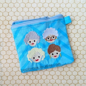 Könnte beinhalten: Hellblaue Reißverschlusstasche mit gestickten Gesichtern von vier Frauen. Die Tasche hat ein Zickzackmuster und eine kleine blaue Lasche. Der Text "designsbyLittleBee.com" ist unten aufgedruckt.