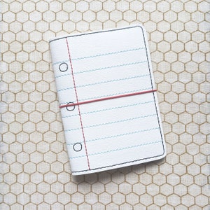 May include: A white notebook cover designed to look like a sheet of lined paper, complete with a red margin line and three black circles. A red elastic band secures the notebook. The cover is on a beige honeycomb pattern.