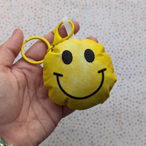 Peut inclure: Un porte-clés jaune en forme de smiley. Le porte-clés est en tissu et présente une bouche souriante noire et deux yeux noirs. Il est équipé d'un anneau et d'un fermoir en métal jaune.