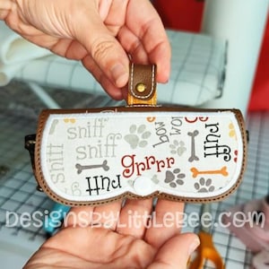 Op de afbeelding: Een brillenkoker met hondenthema met een bruine leren riem en een witte drukknoopsluiting. De koker heeft een witte achtergrond met pootafdrukken, botten en de woorden "sniff", "grrrr" en "puff".