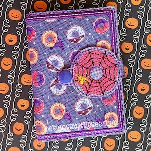 Puede incluir: Una cartera de tela morada con un estampado repetido de donuts y murciélagos. La solapa delantera presenta un diseño de telaraña rosa con una pequeña araña amarilla y un cierre de botón azul. Los bordes están ribeteados con costuras moradas.