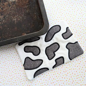 Puede incluir: Un posavasos de algodón blanco con un diseño de vaca negra. El posavasos está hecho de una tela suave y texturizada y tiene un borde negro cosido.