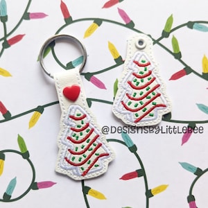 May include: White leather keychain with a red heart and an embroidered Christmas tree design. The tree is decorated with green and red stripes and white dots. The keychain is on a background of colorful Christmas lights.