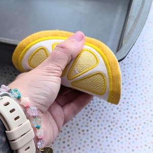 May include: A lemon-shaped pot holder with yellow and white embroidered details. The pot holder is held in a hand, and the background includes a baking pan and a dotted surface.