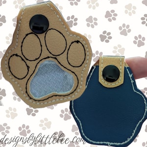 May include: A tan and navy blue paw-shaped keyring with a black snap closure. The tan paw print has black stitching and a mesh centre. The keyring is designed to resemble a dog's paw print. The website "designsbylittlebee.com" is visible in the image.