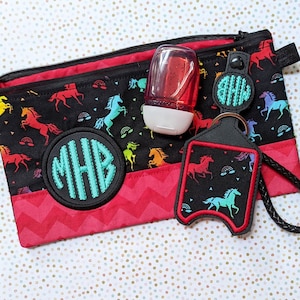 May include: Black zippered pouch with a red chevron accent and a black and red trim. The pouch features a colorful horse pattern and a teal monogram. A matching keychain and hand sanitizer bottle are also included.