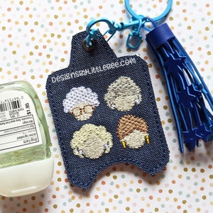 May include: A blue denim hand sanitizer holder with a blue tassel keychain. The holder has embroidered images of four women with blonde hair. The text "DesignSBYLittleBee.com" is printed on the holder.