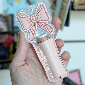 May include: A light pink lip balm holder with a bow design and glitter accents. The holder has a pocket for the lip balm and the text "designsbylittlebee.com" is printed on the side.