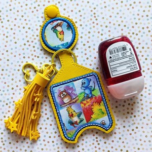 May include: A yellow hand sanitizer holder with a cartoon animal design, a yellow tassel keychain, and a red hand sanitizer bottle. The holder features a blue and white border and a cartoon owl, parrot, and blue mouse.