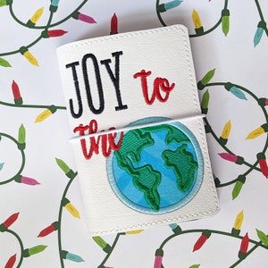 Könnte beinhalten: Ein weißes Ledernotizbuch mit einem weißen Gummibandverschluss. Der Umschlag zeigt einen gestickten Text, der "JOY to the" in roter und schwarzer Farbe lautet. Eine gestickte blaue und grüne Weltkugel befindet sich mittig auf dem Umschlag.