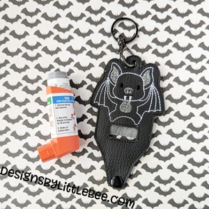 Puede incluir: Un estuche negro de polipiel para inhalador con diseño de murciélago, un llavero negro y un inhalador naranja. El murciélago está bordado con hilo blanco. El estuche tiene un cierre a presión. El fondo es un patrón de murciélagos grises.