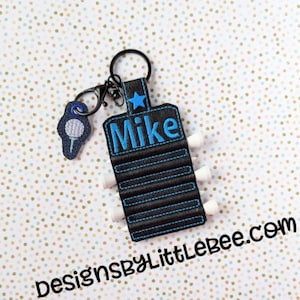 Puede incluir: Un porta tees de golf negro con una estrella azul y el nombre "Mike" bordado. El porta tees tiene seis ranuras para tees de golf y está unido a un llavero plateado con un dije de pelota de golf.